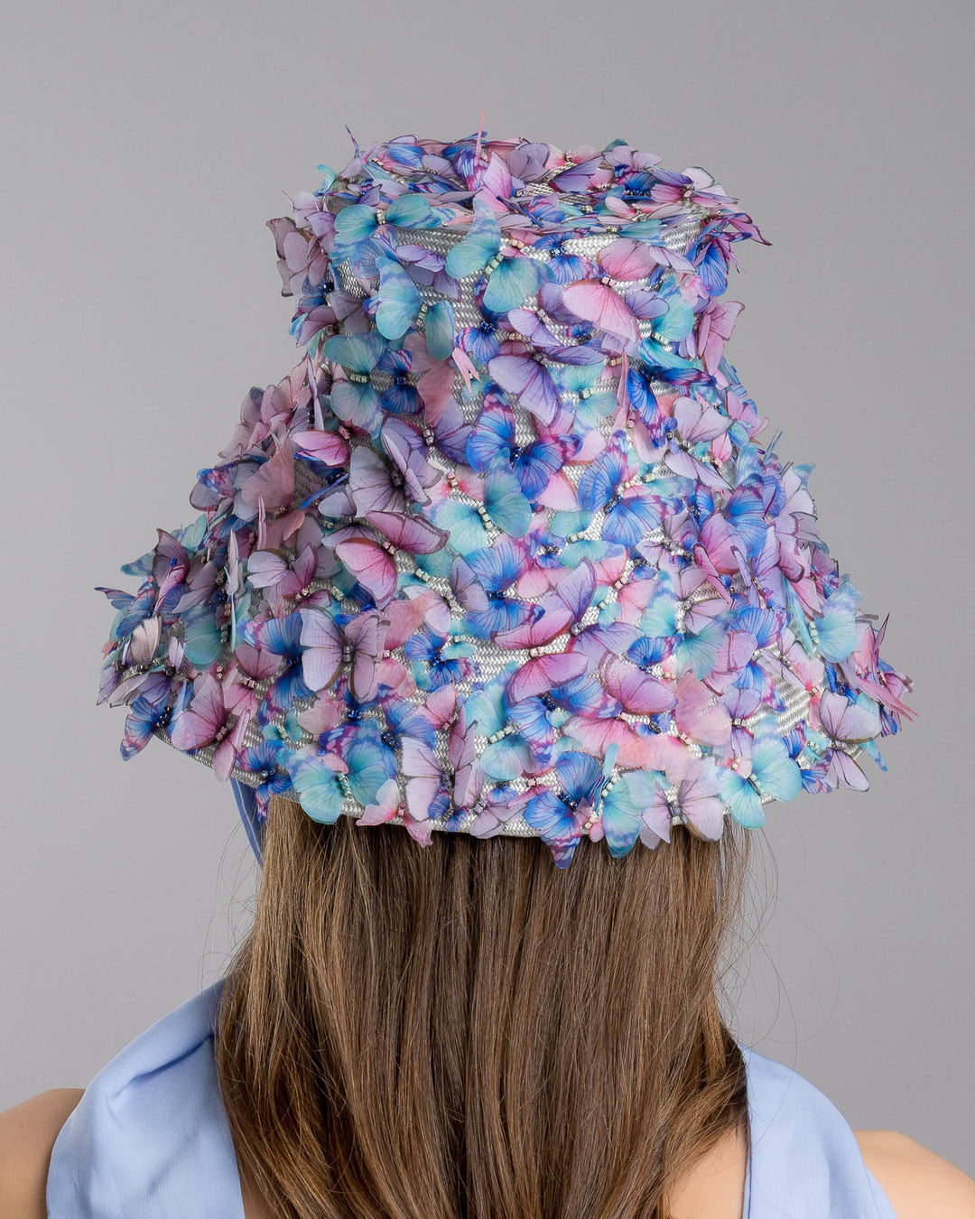 The model showcases the back side of the   high fashion blue bucket hat  with long blue silk chiffon ribbons dropping on the sides, the hat is adorned with multi colour organza butterflies. Designed and made by Merve Bayindir Millinery.