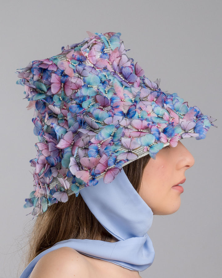 The model showcases the right side of the   high fashion blue bucket hat  with long blue silk chiffon ribbons dropping on the sides, the hat is adorned with multi colour organza butterflies. Designed and made by Merve Bayindir Millinery.