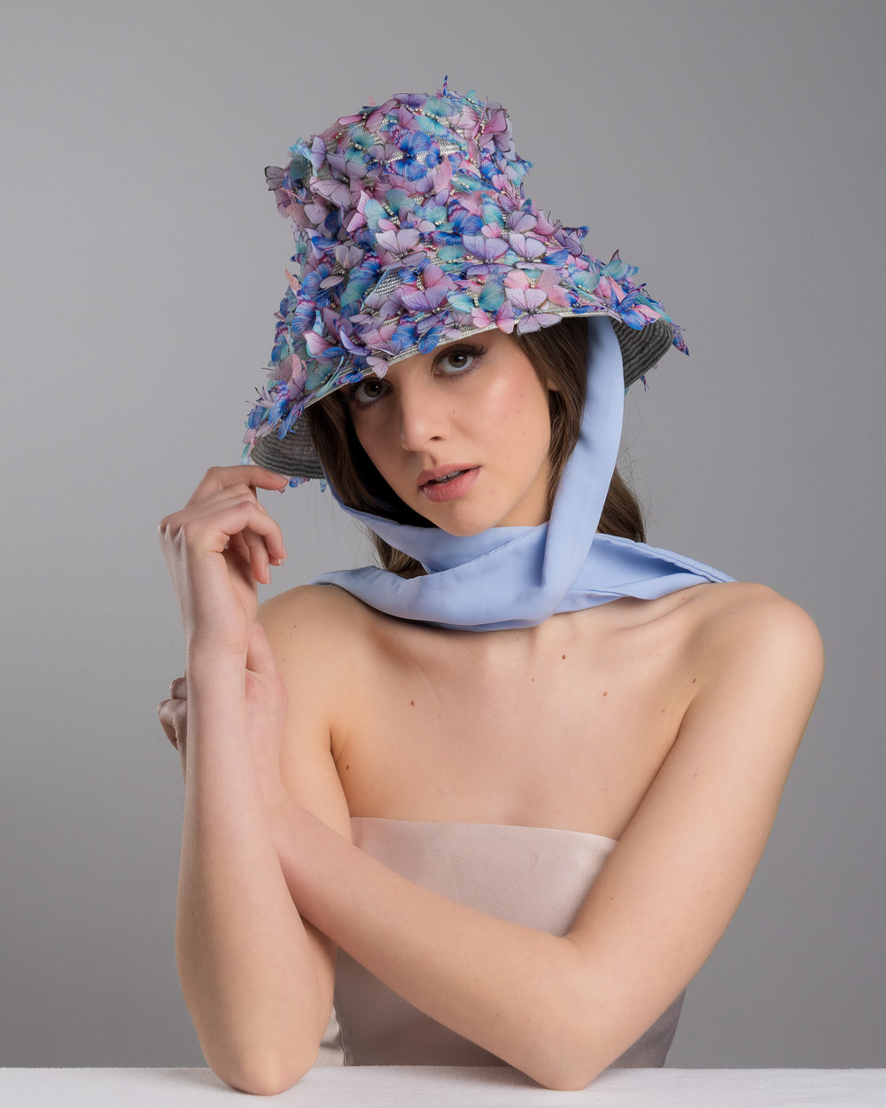The female model is posing, wearing the Butterfly,  high fashion blue bucket hat  with long blue silk chiffon ribbons dropping on the sides, the hat is adorned with multi colour organza butterflies. Designed and made by Merve Bayindir Millinery.