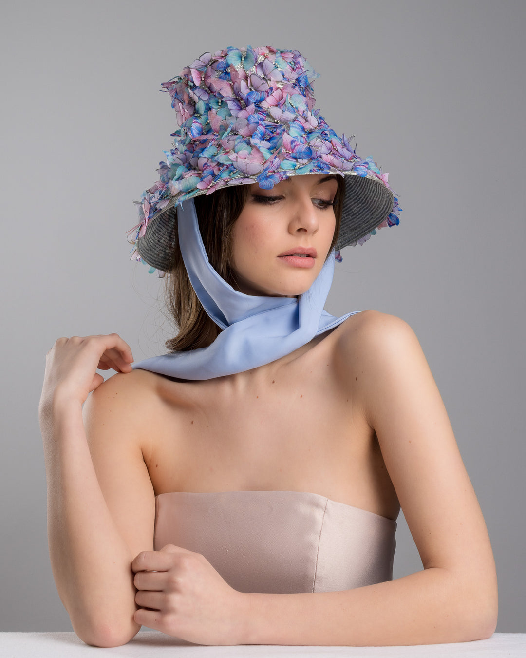 The female model is posing, wearing the Butterfly,  high fashion blue bucket hat  with long blue silk chiffon ribbons dropping on the sides, the hat is adorned with multi colour organza butterflies. Designed and made by Merve Bayindir Millinery.