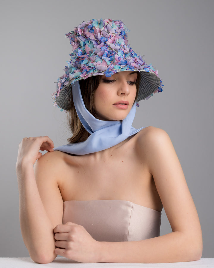 The female model is posing, wearing the Butterfly,  high fashion blue bucket hat  with long blue silk chiffon ribbons dropping on the sides, the hat is adorned with multi colour organza butterflies. Designed and made by Merve Bayindir Millinery.