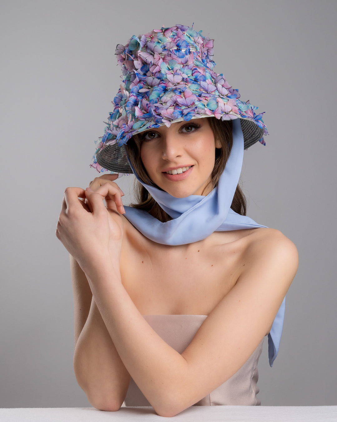 The female model is posing, wearing the Butterfly,  high fashion blue bucket hat  with long blue silk chiffon ribbons dropping on the sides, the hat is adorned with multi colour organza butterflies. Designed and made by Merve Bayindir Millinery.