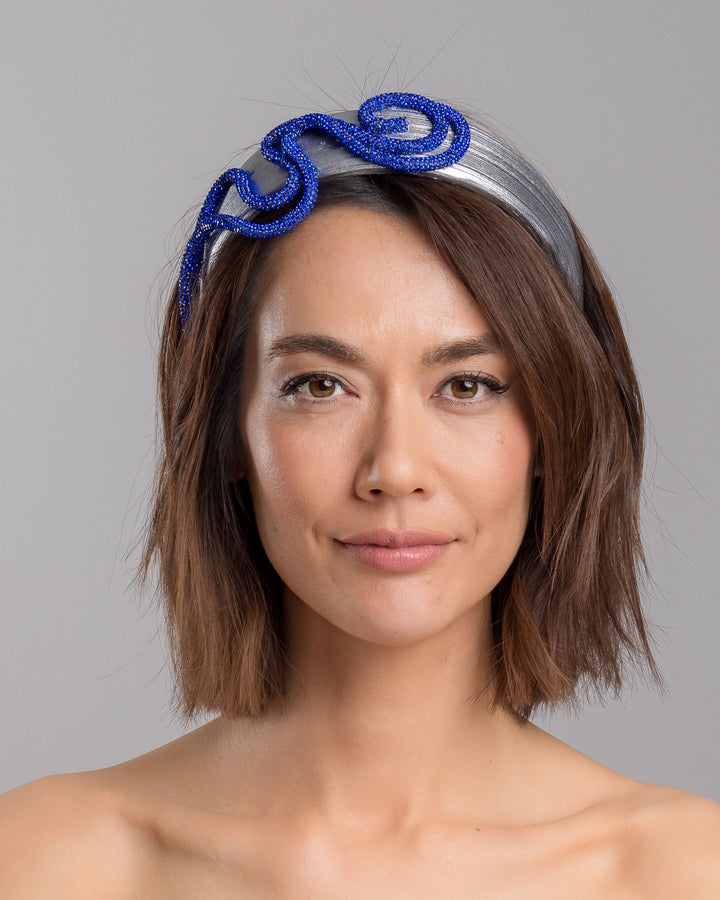 The model is showcasing the front side close up of the light silver headband with a faux leather base, wrapped in bold looping cobalt blue crystal-embellished cords, creating a sculptural, high-shine statement design. Designed and made by Merve Bayindir Millinery.
