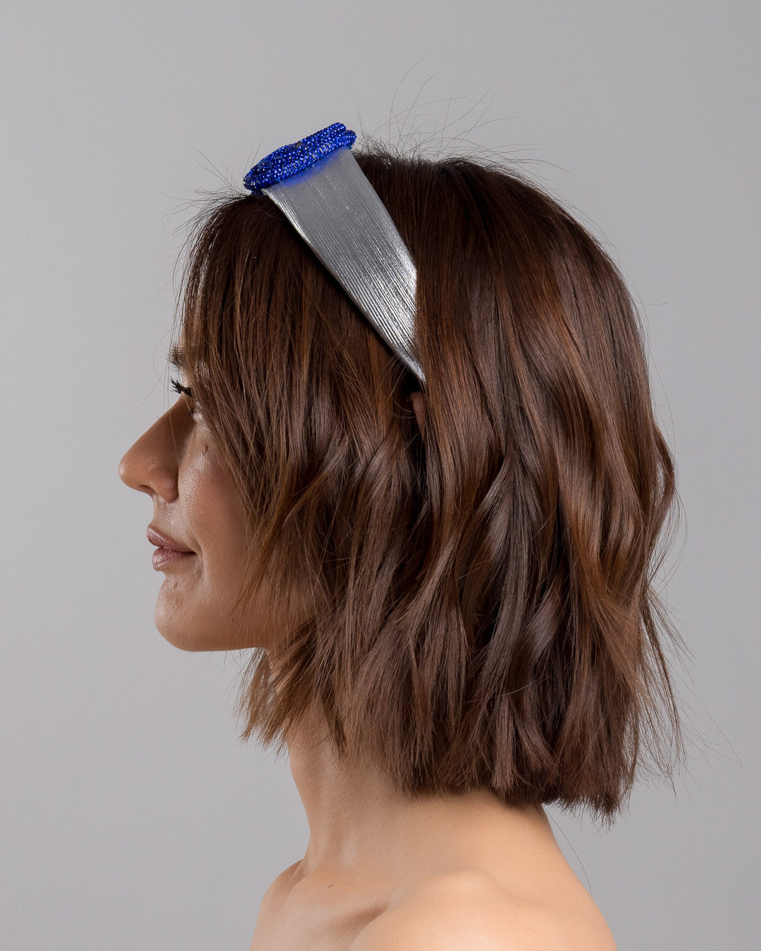 The model is showcasing the left side close up of the light silver headband with a faux leather base, wrapped in bold looping cobalt blue crystal-embellished cords, creating a sculptural, high-shine statement design. Designed and made by Merve Bayindir Millinery.
