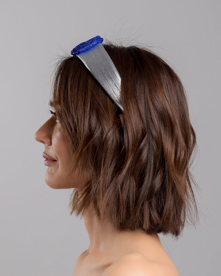 The model is showcasing the left side close up of the light silver headband with a faux leather base, wrapped in bold looping cobalt blue crystal-embellished cords, creating a sculptural, high-shine statement design. Designed and made by Merve Bayindir Millinery.
