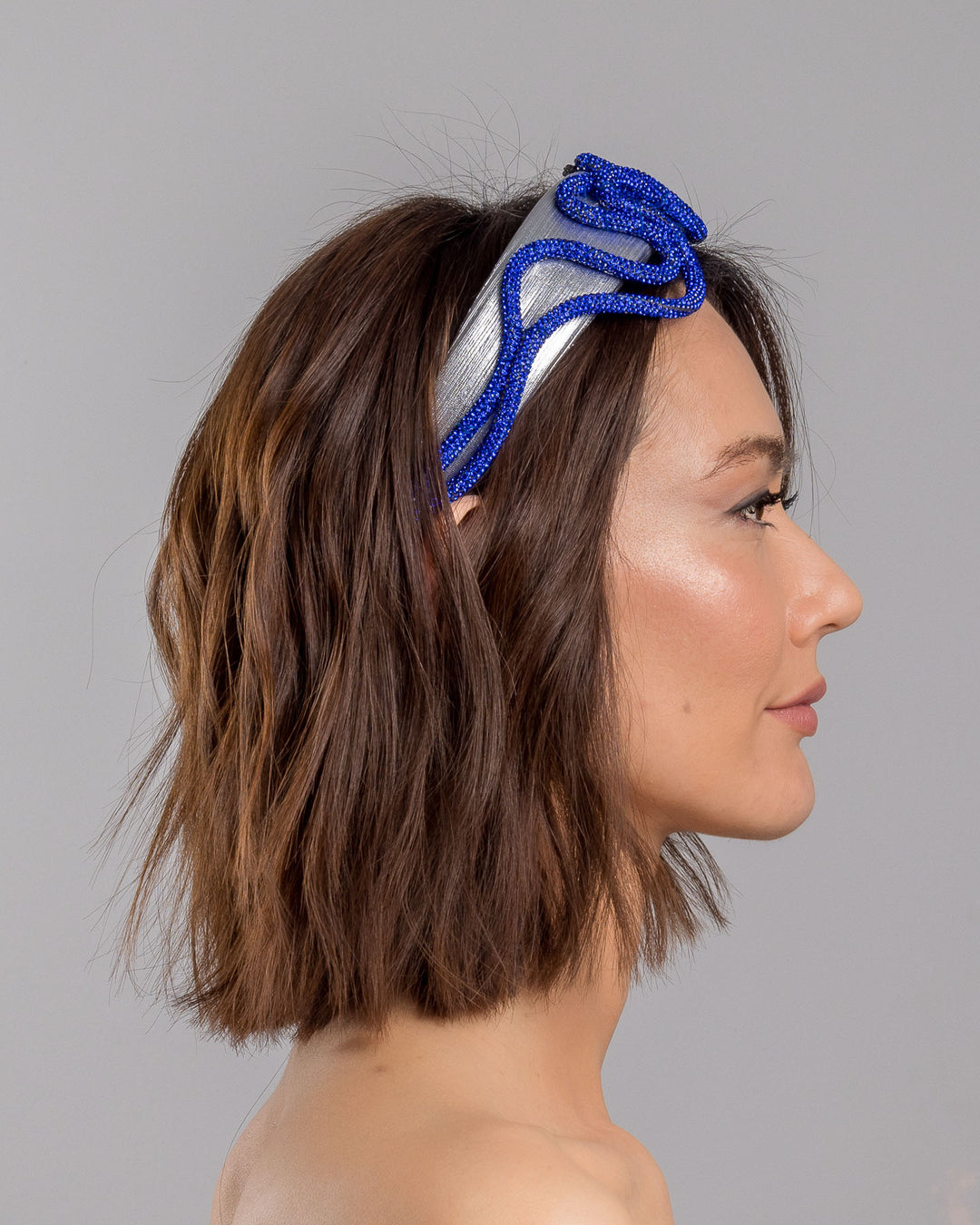 The model is showcasing the right side close up of the light silver headband with a faux leather base, wrapped in bold looping cobalt blue crystal-embellished cords, creating a sculptural, high-shine statement design. Designed and made by Merve Bayindir Millinery.

