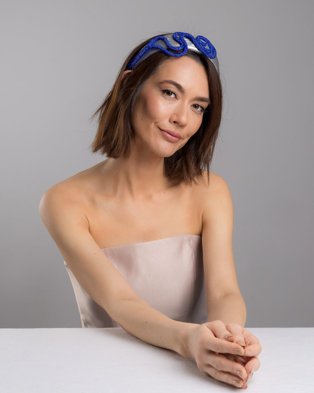 The female model is posing, wearing Cereal, the light silver headband with a faux leather base, wrapped in bold looping cobalt blue crystal-embellished cords, creating a sculptural, high-shine statement design. Designed and made by Merve Bayindir Millinery.
