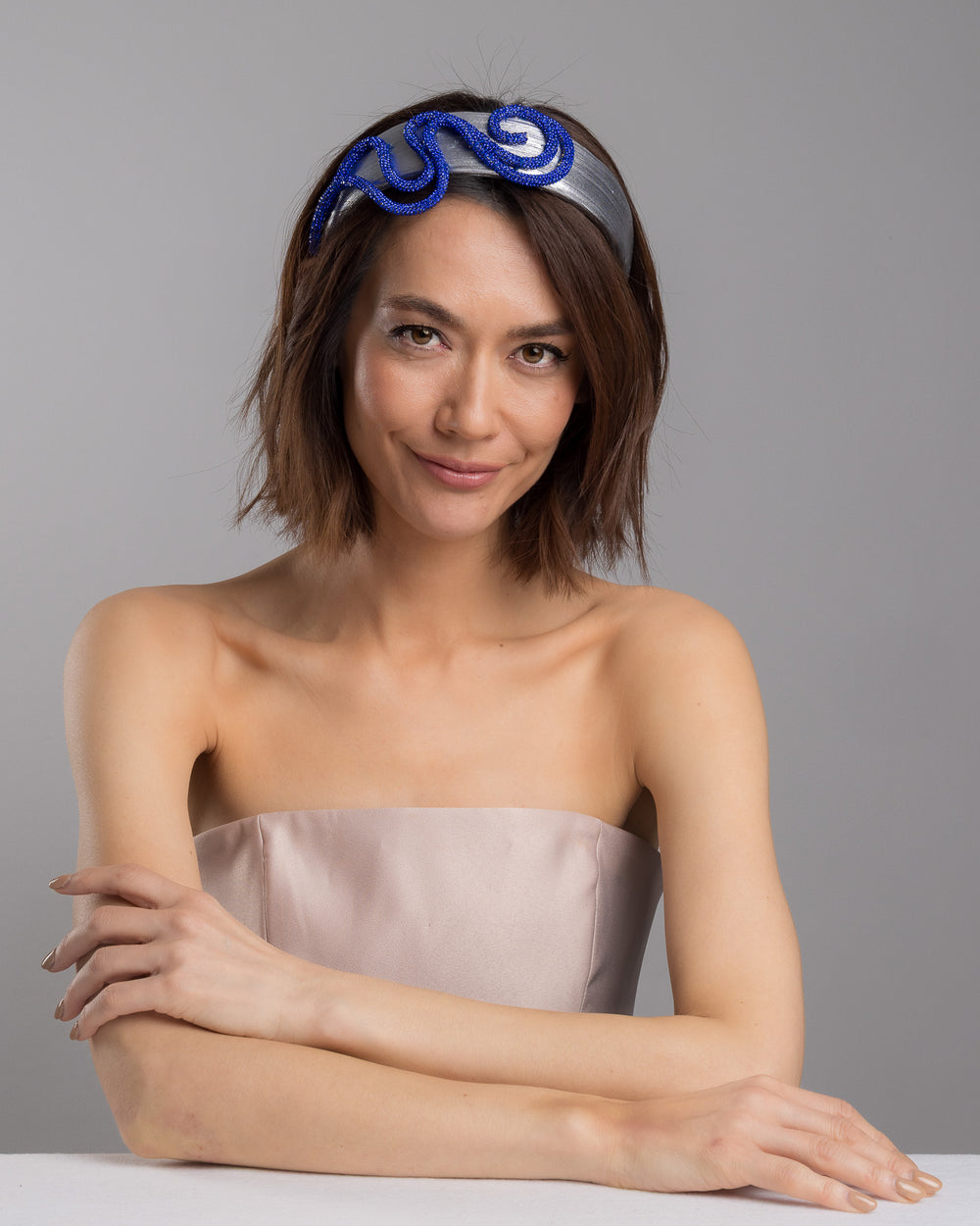 The female model is posing, wearing Cereal, the light silver headband with a faux leather base, wrapped in bold looping cobalt blue crystal-embellished cords, creating a sculptural, high-shine statement design. Designed and made by Merve Bayindir Millinery.
