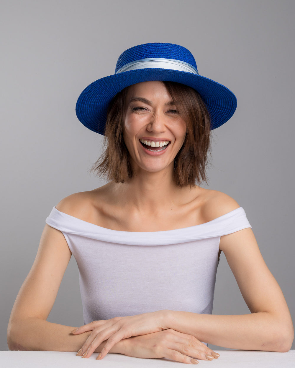 The female model is posing, wearing Cielor, the royal blue woven straw boater hat with a wide brim, wrapped in a soft pale grey silk ribbon and finished with long scarf ties featuring delicate illustrated figures. Designed and made by Merve Bayindir millinery.