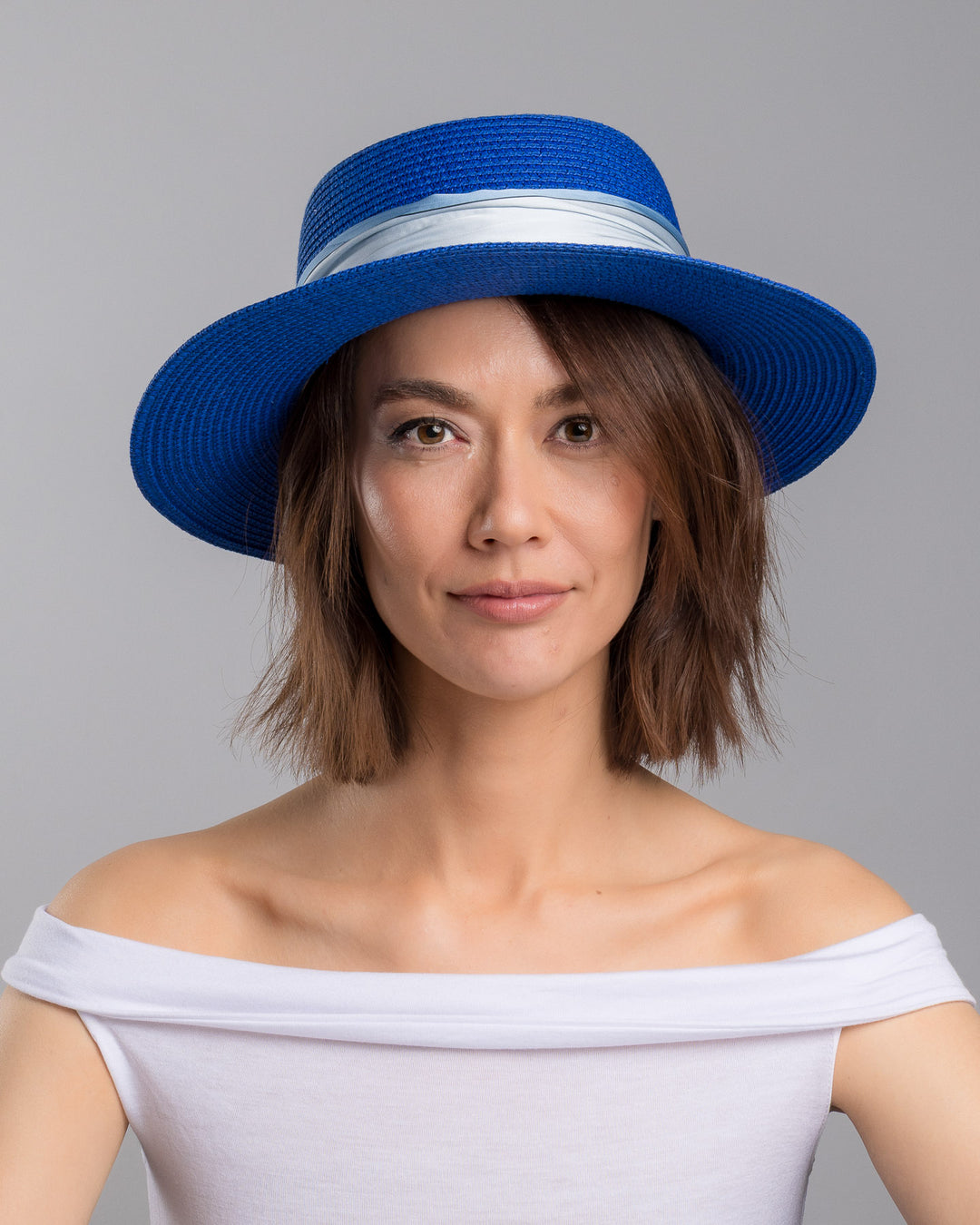 The model is showcasing the front side close up of the royal blue woven straw boater hat with a wide brim, wrapped in a soft pale grey silk ribbon and finished with long scarf ties featuring delicate illustrated figures. Designed and made by Merve Bayindir millinery.