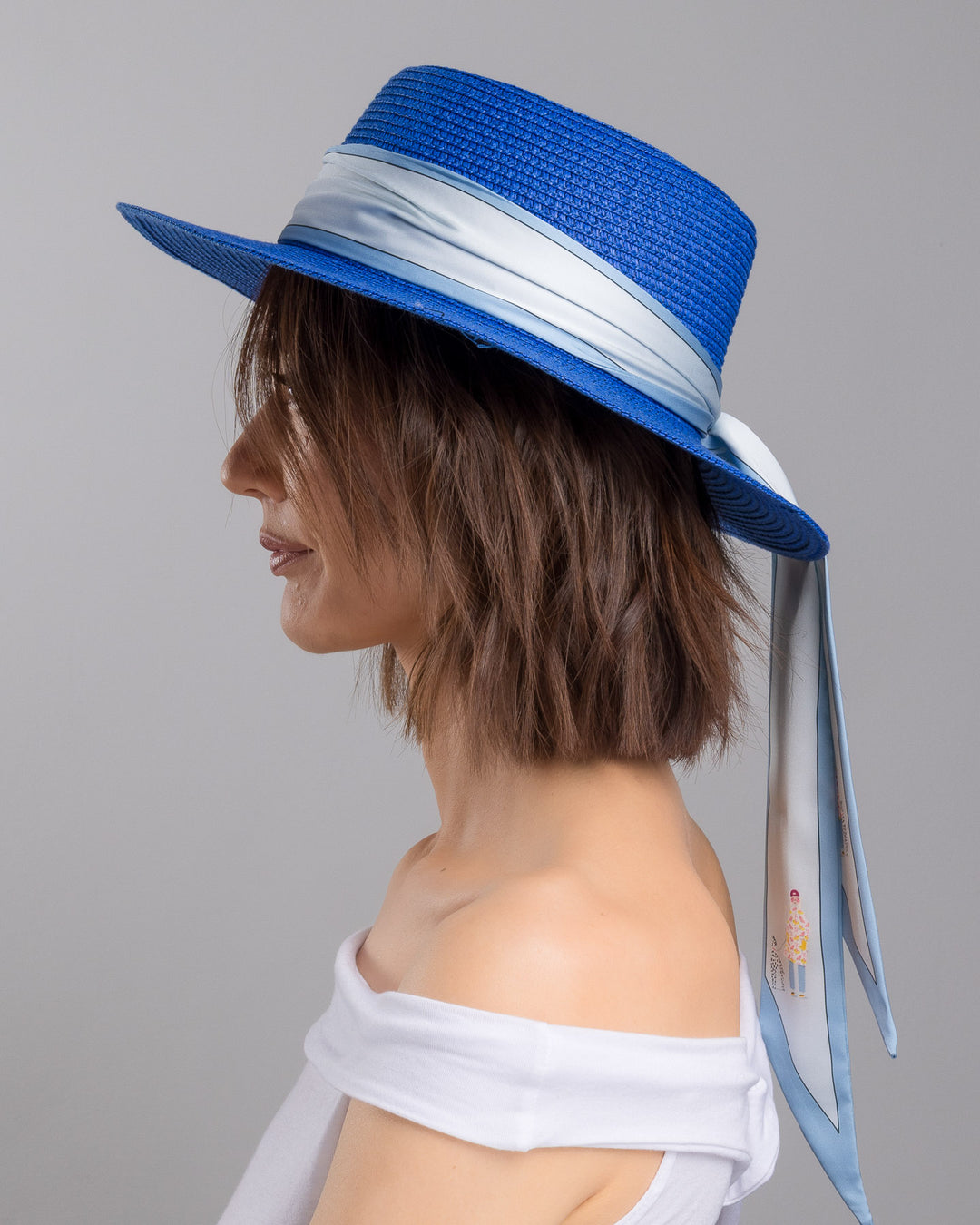 The model is showcasing the left side close up of the royal blue woven straw boater hat with a wide brim, wrapped in a soft pale grey silk ribbon and finished with long scarf ties featuring delicate illustrated figures. Designed and made by Merve Bayindir millinery.