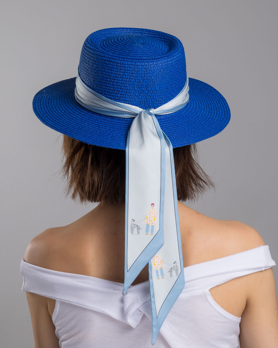 The model is showcasing the back side close up of the royal blue woven straw boater hat with a wide brim, wrapped in a soft pale grey silk ribbon and finished with long scarf ties featuring delicate illustrated figures. Designed and made by Merve Bayindir millinery.