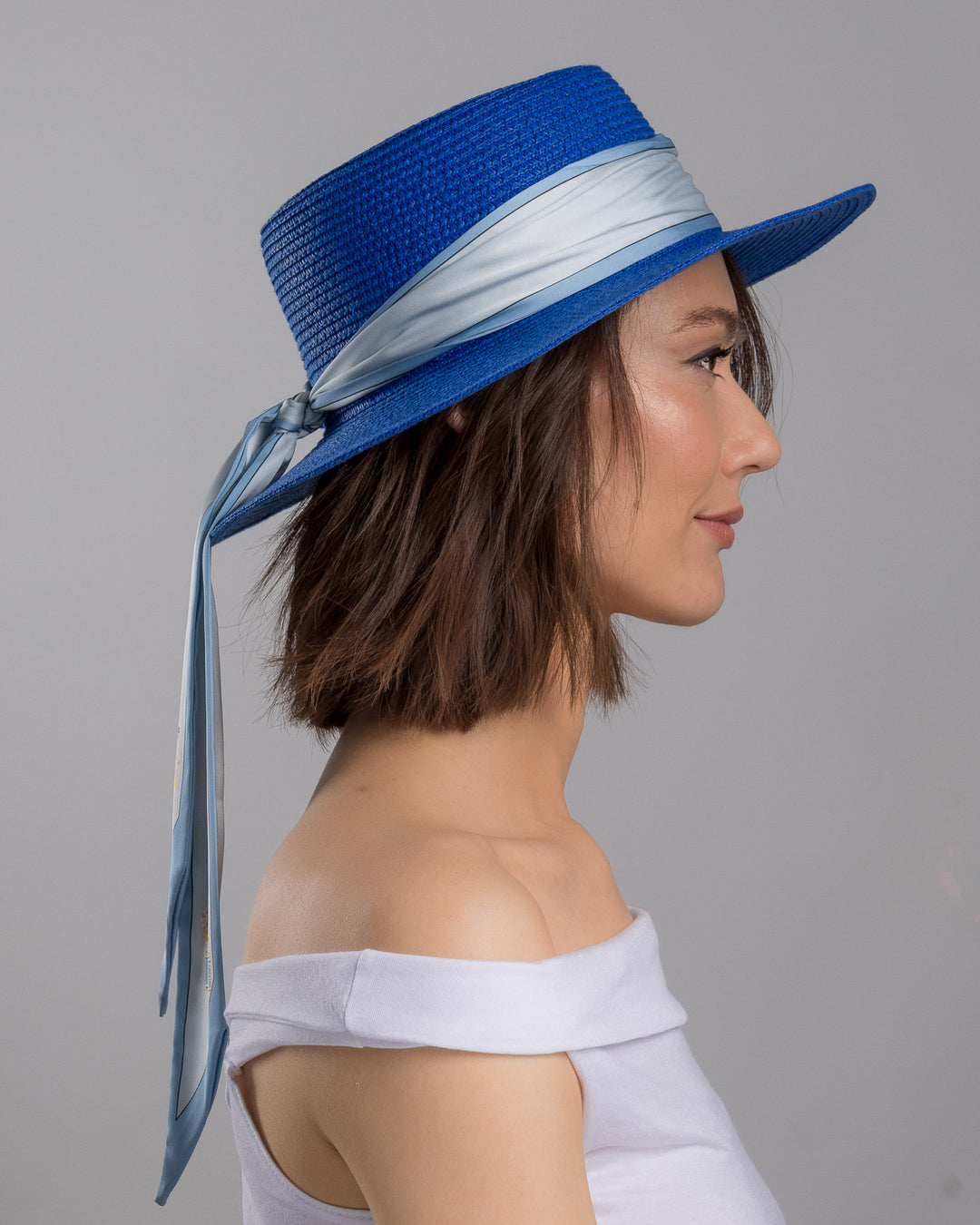 The model is showcasing the right side close up of the royal blue woven straw boater hat with a wide brim, wrapped in a soft pale grey silk ribbon and finished with long scarf ties featuring delicate illustrated figures. Designed and made by Merve Bayindir millinery.