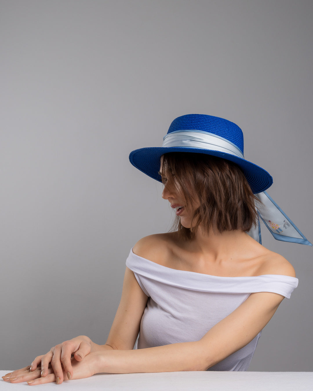 The female model is posing, wearing Cielor, the royal blue woven straw boater hat with a wide brim, wrapped in a soft pale grey silk ribbon and finished with long scarf ties featuring delicate illustrated figures. Designed and made by Merve Bayindir millinery.