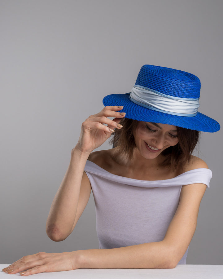 The female model is posing, wearing Cielor, the royal blue woven straw boater hat with a wide brim, wrapped in a soft pale grey silk ribbon and finished with long scarf ties featuring delicate illustrated figures. Designed and made by Merve Bayindir millinery.