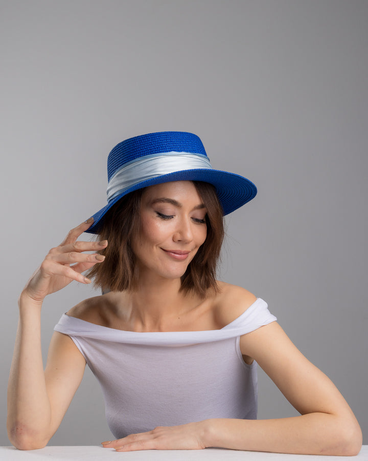 The female model is posing, wearing Cielor, the royal blue woven straw boater hat with a wide brim, wrapped in a soft pale grey silk ribbon and finished with long scarf ties featuring delicate illustrated figures. Designed and made by Merve Bayindir millinery.