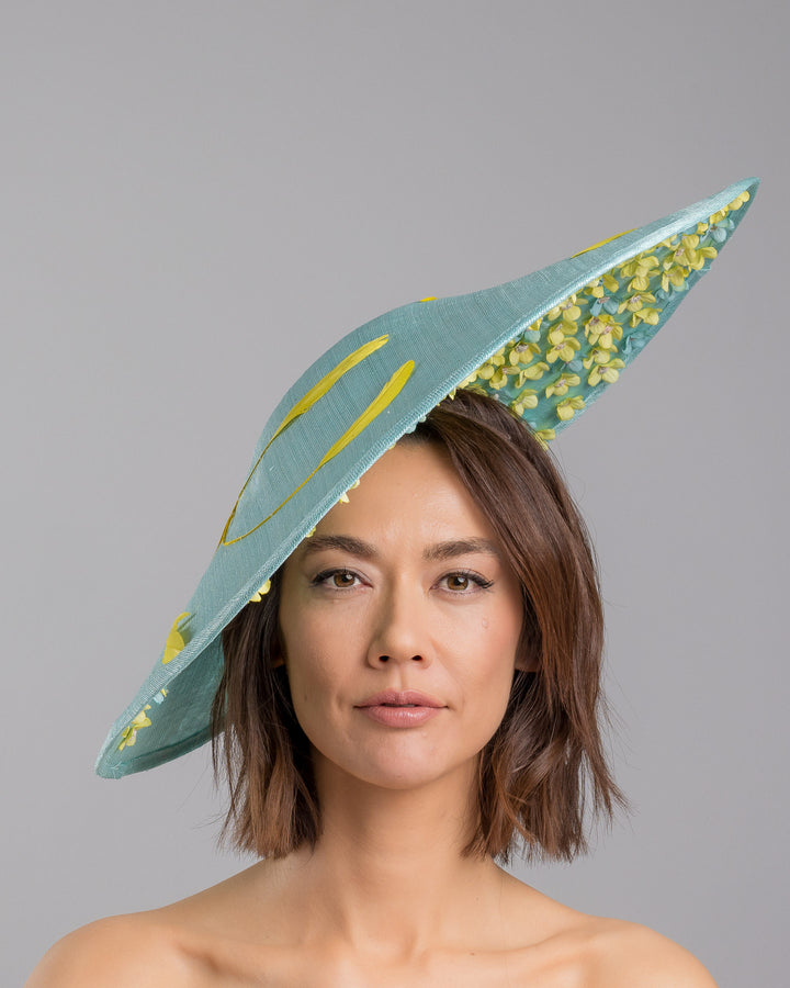 The model is showcasing the front side close up of the mint coloured sinamay base hat with a wide sculpted brim, featuring delicate yellow flowers lining the underside and finished with a bold yellow feather accent on the outer edge for a striking contrast. Designed and made by Merve Bayindir Millinery.
