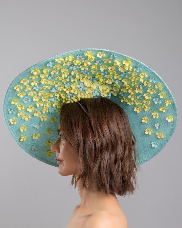 The model is showcasing the left side close up of the mint coloured sinamay base hat with a wide sculpted brim, featuring delicate yellow flowers lining the underside and finished with a bold yellow feather accent on the outer edge for a striking contrast. Designed and made by Merve Bayindir Millinery.
