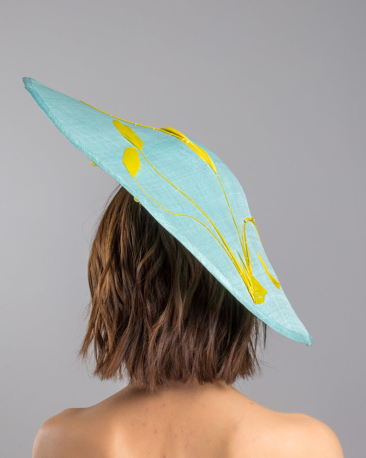 The model is showcasing the back side close up of the mint coloured sinamay base hat with a wide sculpted brim, featuring delicate yellow flowers lining the underside and finished with a bold yellow feather accent on the outer edge for a striking contrast. Designed and made by Merve Bayindir Millinery.
