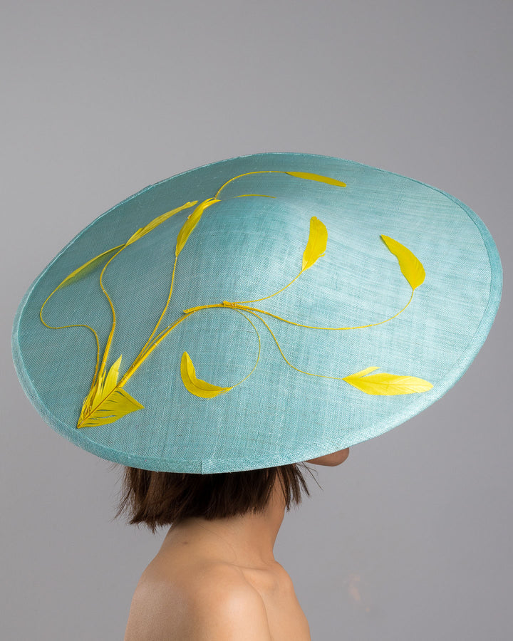 The model is showcasing the right side close up of the mint coloured sinamay base hat with a wide sculpted brim, featuring delicate yellow flowers lining the underside and finished with a bold yellow feather accent on the outer edge for a striking contrast. Designed and made by Merve Bayindir Millinery.
