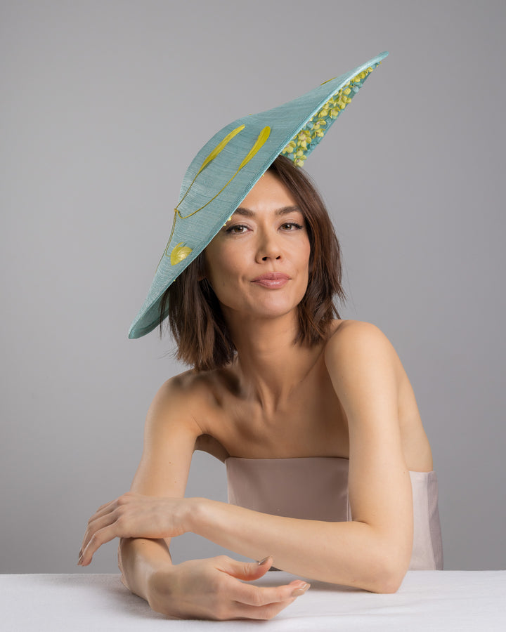 The female model is posing, wearing
Clyra, the mint coloured sinamay base hat with a wide sculpted brim, featuring delicate yellow flowers lining the underside and finished with a bold yellow feather accent on the outer edge for a striking contrast. Designed and made by Merve Bayindir Millinery.
