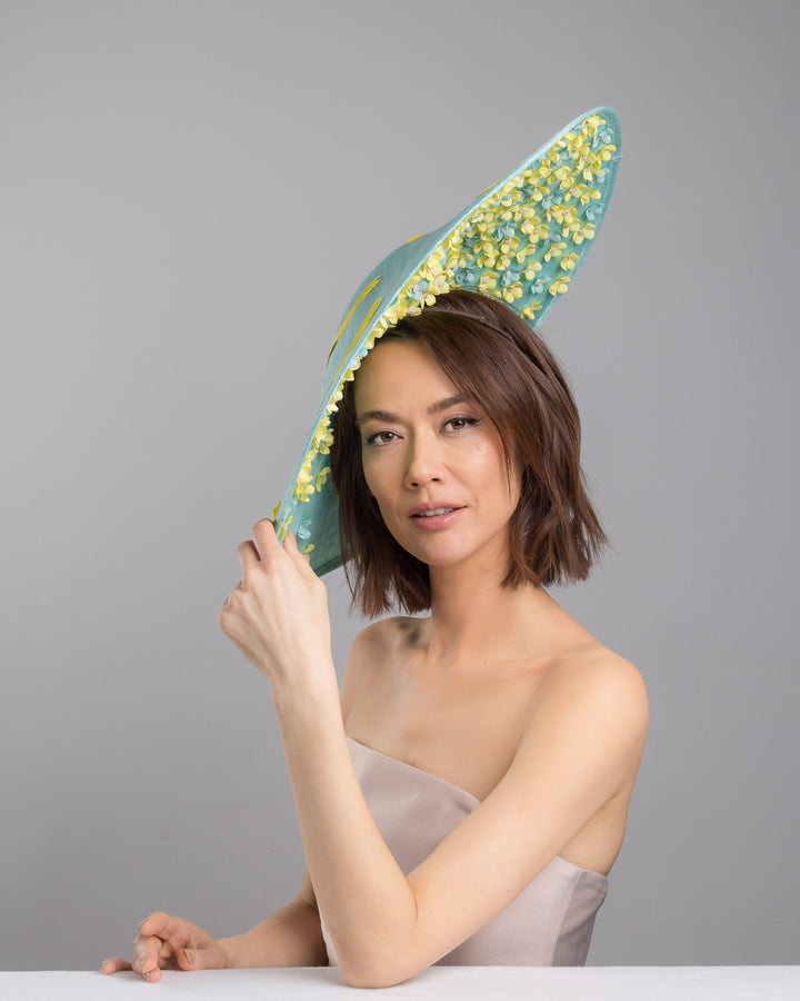 The female model is posing, wearing
Clyra, the mint coloured sinamay base hat with a wide sculpted brim, featuring delicate yellow flowers lining the underside and finished with a bold yellow feather accent on the outer edge for a striking contrast. Designed and made by Merve Bayindir Millinery.
