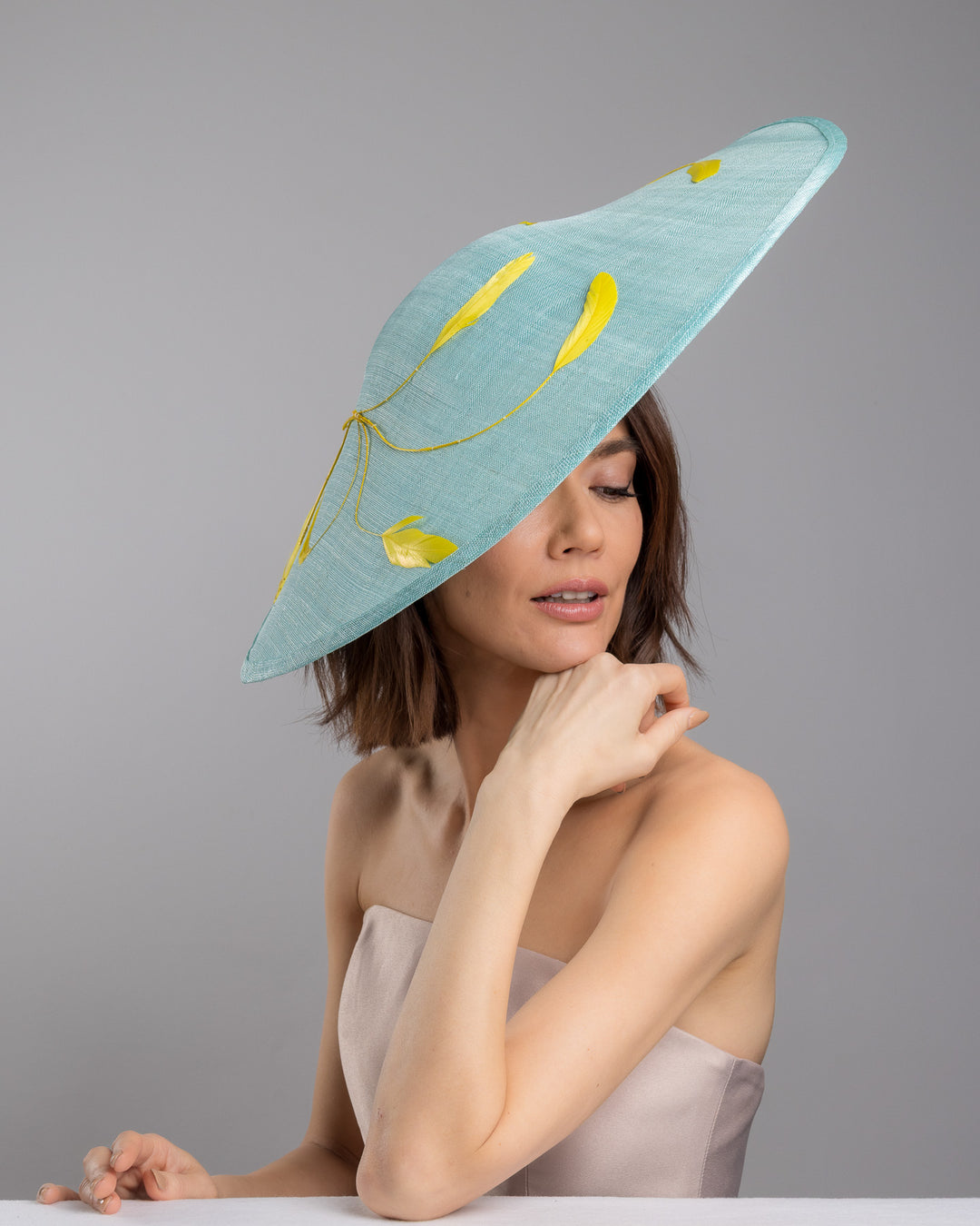 The female model is posing, wearing
Clyra, the mint coloured sinamay base hat with a wide sculpted brim, featuring delicate yellow flowers lining the underside and finished with a bold yellow feather accent on the outer edge for a striking contrast. Designed and made by Merve Bayindir Millinery.
