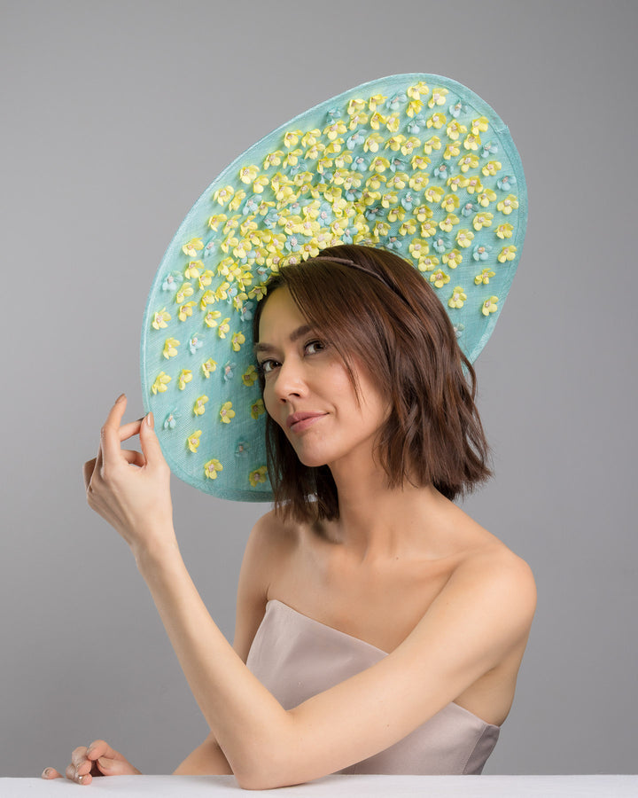 The female model is posing, wearing
Clyra, the mint coloured sinamay base hat with a wide sculpted brim, featuring delicate yellow flowers lining the underside and finished with a bold yellow feather accent on the outer edge for a striking contrast. Designed and made by Merve Bayindir Millinery.
