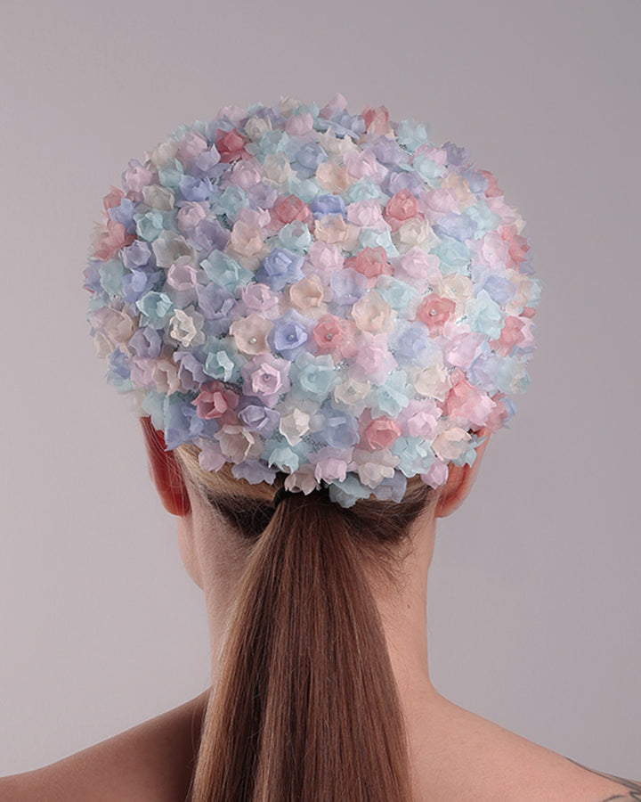 The model is presenting the back of the blue pillbox style hat, embellished with multi colour silk organza flowers.
