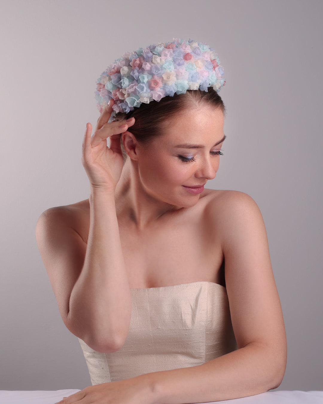 The model is presenting the right top angle of a blue pillbox style hat, embellished with multi colour silk organza flowers.
