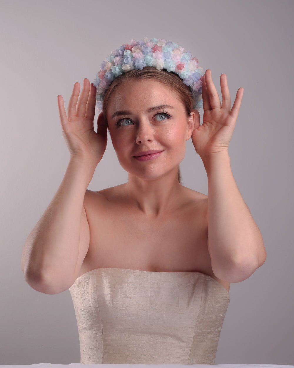 The model is presenting the front angle of a blue pillbox style hat, embellished with multi colour silk organza flowers.