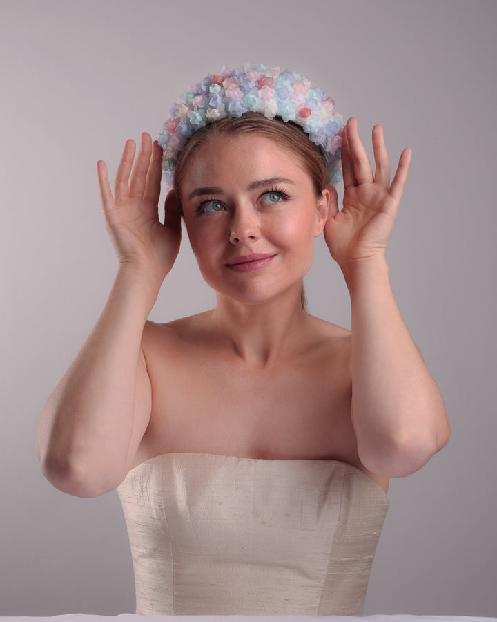The model is presenting the front angle of a blue pillbox style hat, embellished with multi colour silk organza flowers.