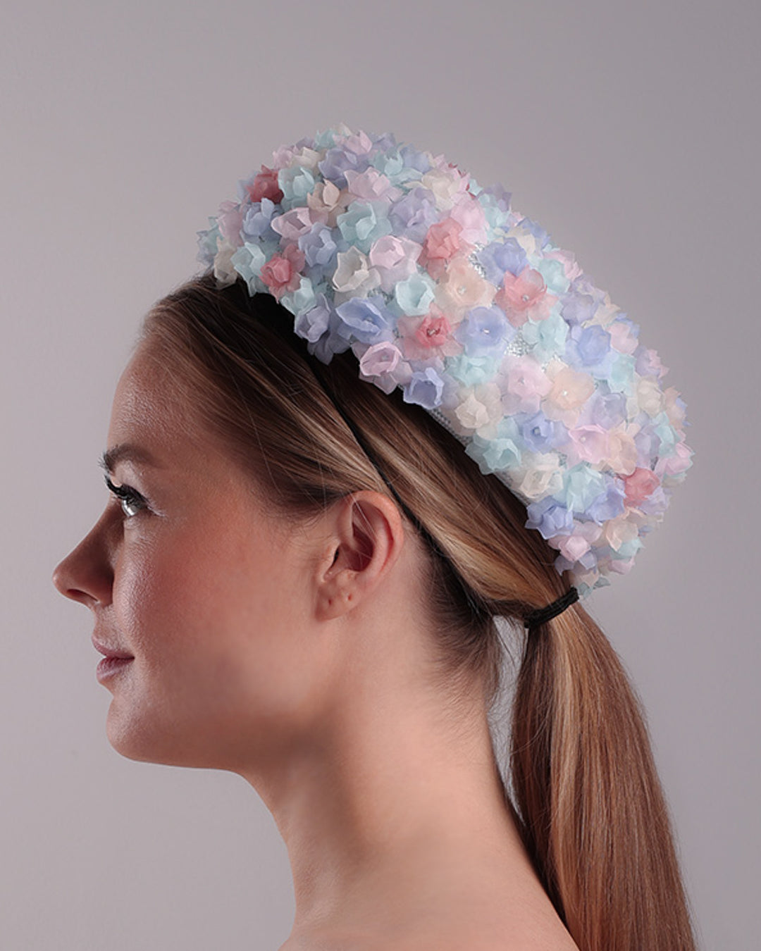 The model is presenting the left side of the blue pillbox style hat, embellished with multi colour silk organza flowers.