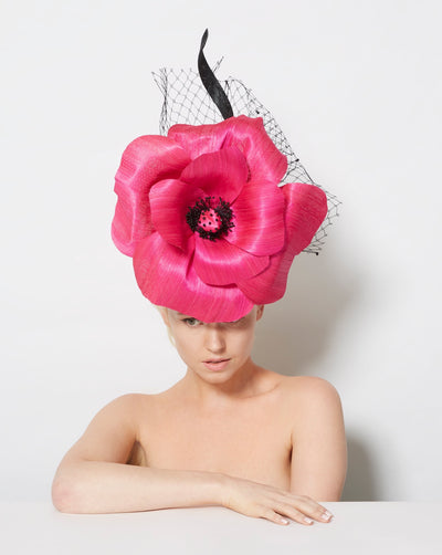 The hot pink fascinator is designed to resemble an oversized, flower. The petals are crafted from a lustrous silk abaca that gives them a realistic yet artistic appearance. At the centre of the flower is a detailed stamen made of small black beads, adding texture and contrast to the design