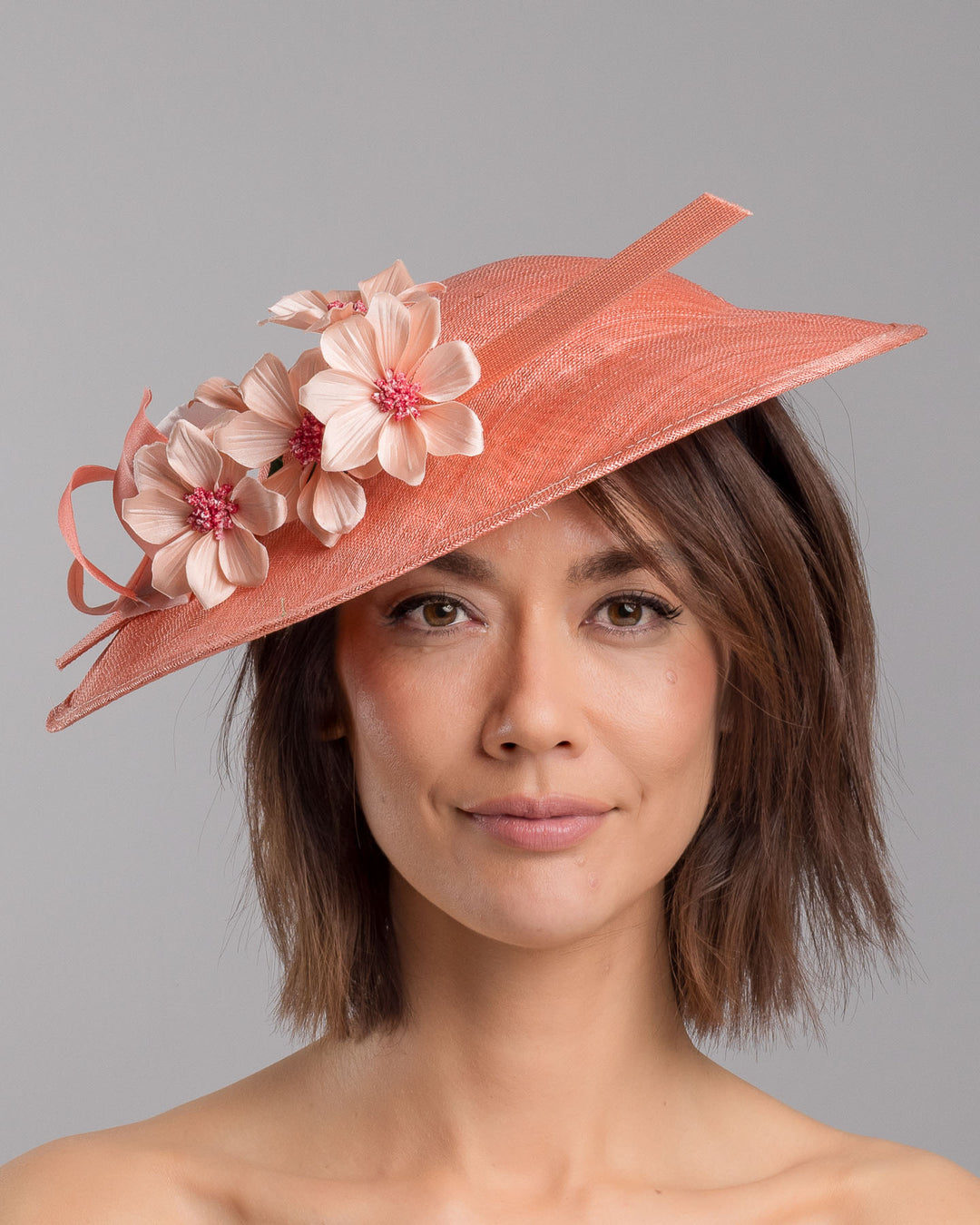 The model is showcasing the front side close up of the coral coloured fascinator hat featuring a wide, asymmetrical brim. The hat is adorned with three large, intricately detailed flowers, each with soft, coral coloured petals and pink beaded centres. Designed and made by Merve Bayindir Millinery.
