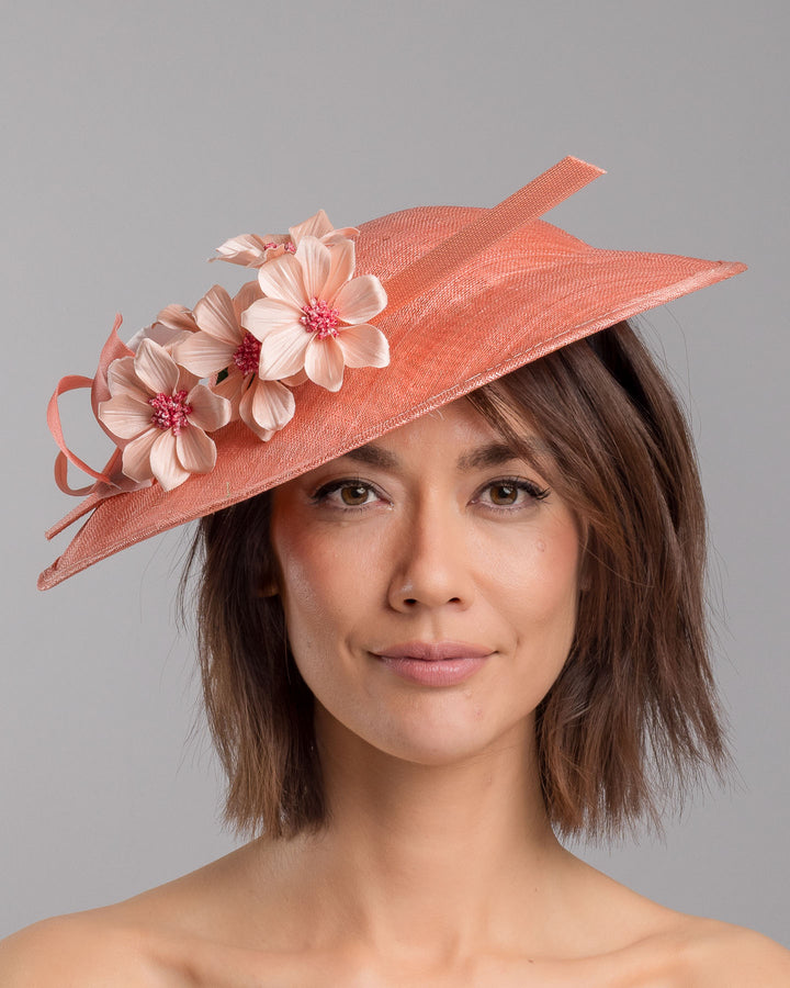 The model is showcasing the front side close up of the coral coloured fascinator hat featuring a wide, asymmetrical brim. The hat is adorned with three large, intricately detailed flowers, each with soft, coral coloured petals and pink beaded centres. Designed and made by Merve Bayindir Millinery.
