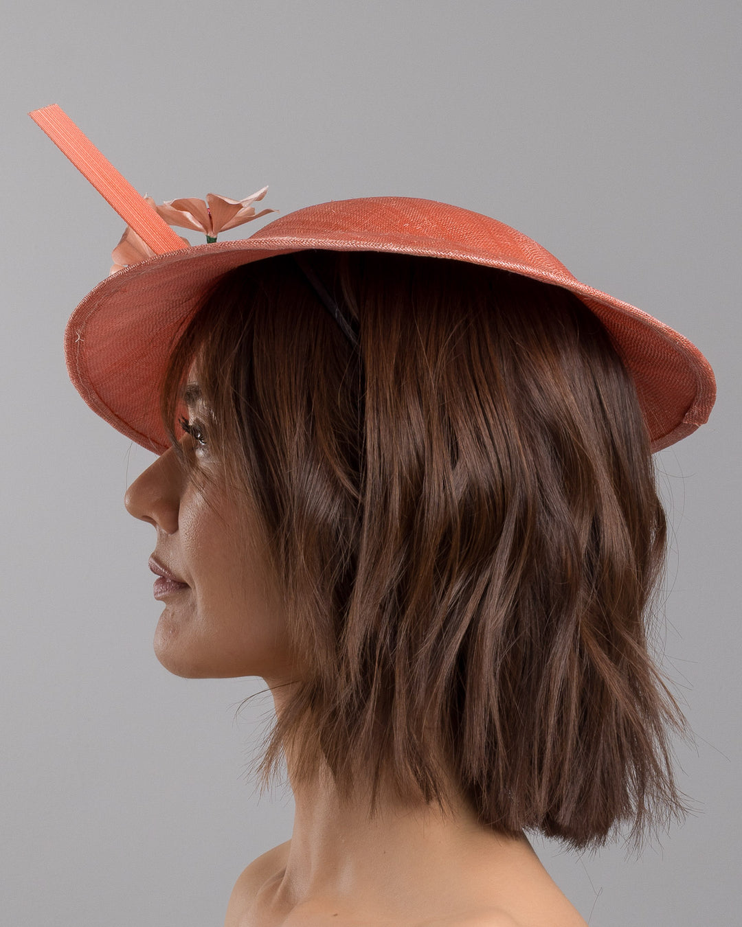 The model is showcasing the left side close up of the coral coloured fascinator hat featuring a wide, asymmetrical brim. The hat is adorned with three large, intricately detailed flowers, each with soft, coral coloured petals and pink beaded centres. Designed and made by Merve Bayindir Millinery.
