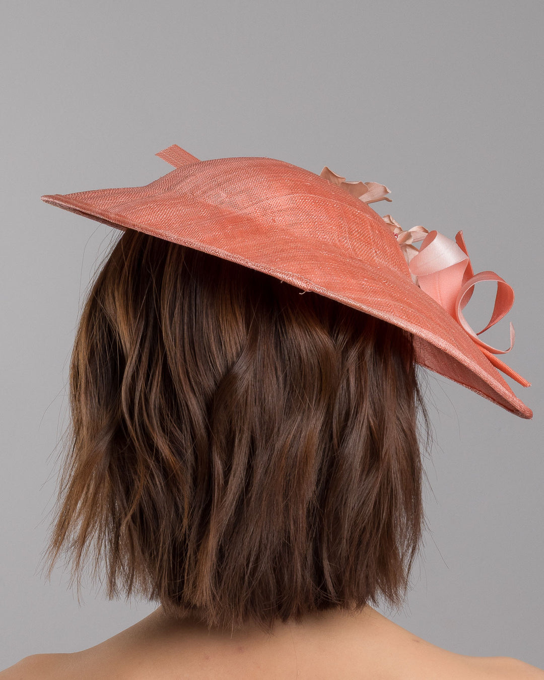 The model is showcasing the back side close up of the coral coloured fascinator hat featuring a wide, asymmetrical brim. The hat is adorned with three large, intricately detailed flowers, each with soft, coral coloured petals and pink beaded centres. Designed and made by Merve Bayindir Millinery.
