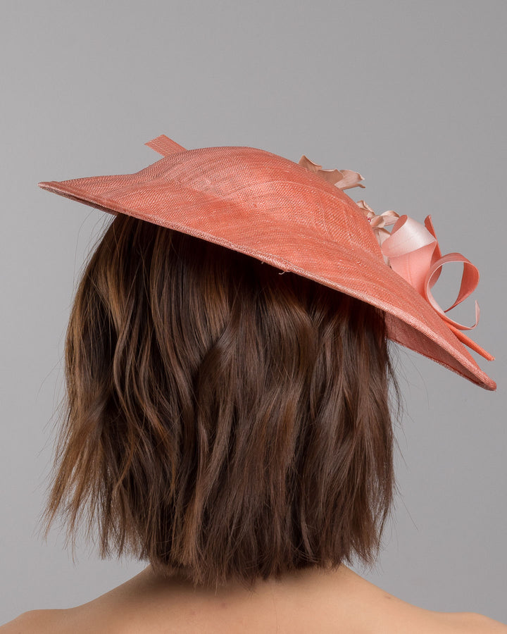 The model is showcasing the back side close up of the coral coloured fascinator hat featuring a wide, asymmetrical brim. The hat is adorned with three large, intricately detailed flowers, each with soft, coral coloured petals and pink beaded centres. Designed and made by Merve Bayindir Millinery.
