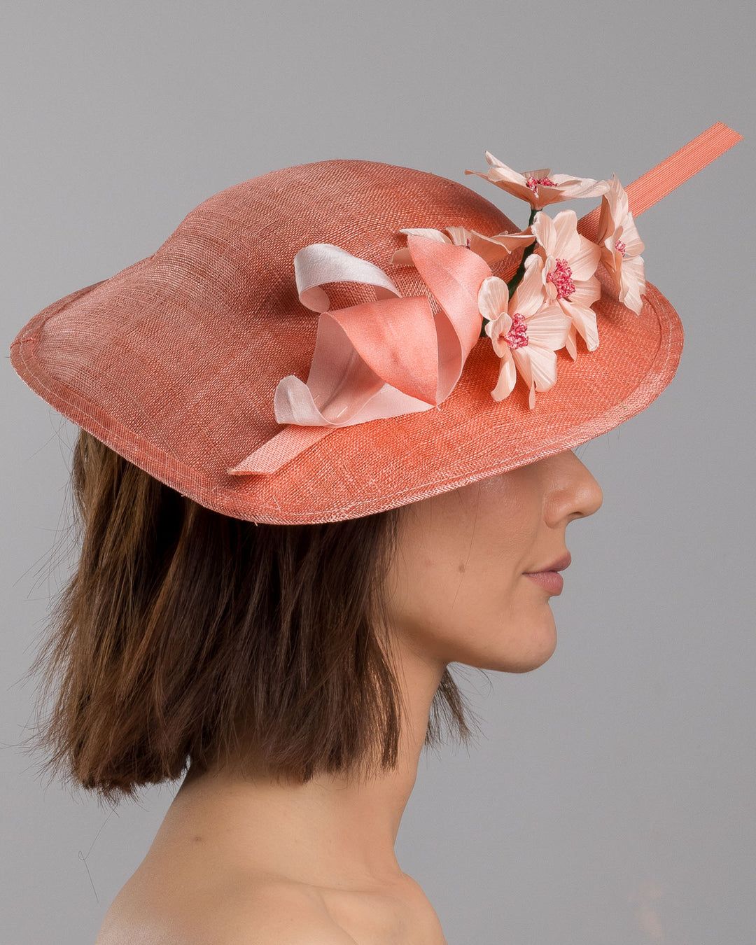 The model is showcasing the right side close up of the coral coloured fascinator hat featuring a wide, asymmetrical brim. The hat is adorned with three large, intricately detailed flowers, each with soft, coral coloured petals and pink beaded centres. Designed and made by Merve Bayindir Millinery.
