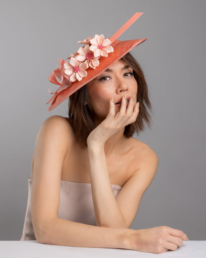The female model is posing, wearing Carry, the coral coloured fascinator hat featuring a wide, asymmetrical brim. The hat is adorned with three large, intricately detailed flowers, each with soft, coral coloured petals and pink beaded centres. Designed and made by Merve Bayindir Millinery.

