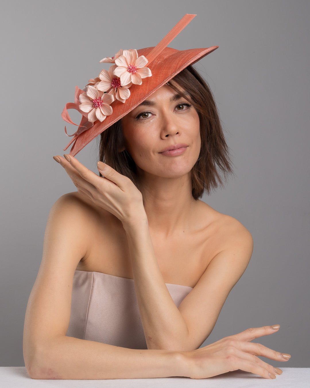The female model is posing, wearing Carry, the coral coloured fascinator hat featuring a wide, asymmetrical brim. The hat is adorned with three large, intricately detailed flowers, each with soft, coral coloured petals and pink beaded centres. Designed and made by Merve Bayindir Millinery.
