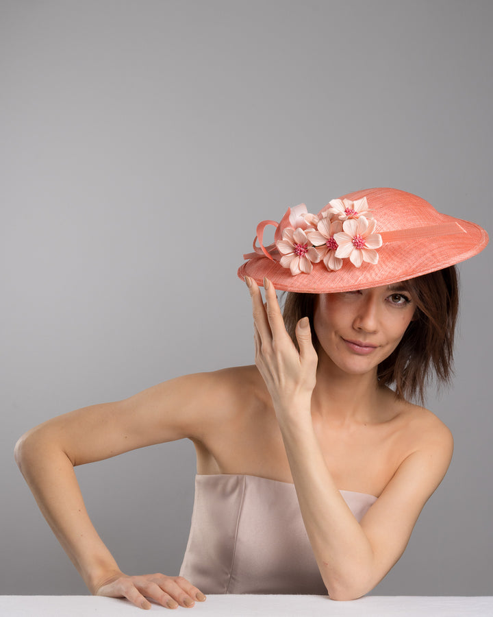 The female model is posing, wearing Carry, the coral coloured fascinator hat featuring a wide, asymmetrical brim. The hat is adorned with three large, intricately detailed flowers, each with soft, coral coloured petals and pink beaded centres. Designed and made by Merve Bayindir Millinery.
