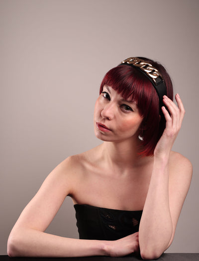 The model showcases the front from the left angle of the Celeste Headband, handcrafted from a satin black Alice headband and crowned with a statement gold chain. Designed by Merve Bayindir.