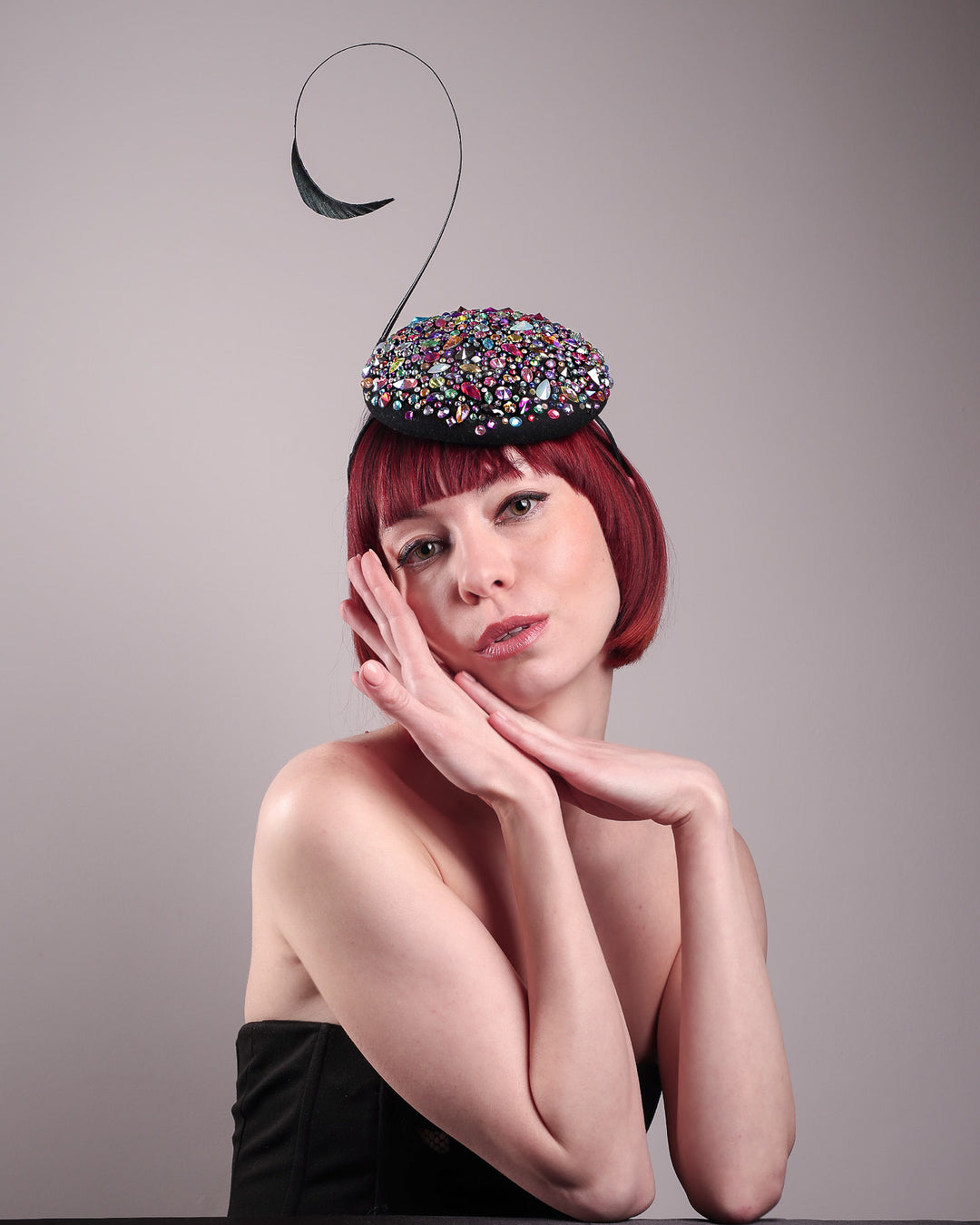 The model showcases the front of the Celestine pillbox fascinator. Its structured, button-shaped black base is drenched in a kaleidoscope of iridescent crystals—pink, aqua, violet, and gold, among others—each catching the light in motion. Crowned with a sweeping black quill that curves like a clef in mid-air. Designed by Merve Bayindir.
