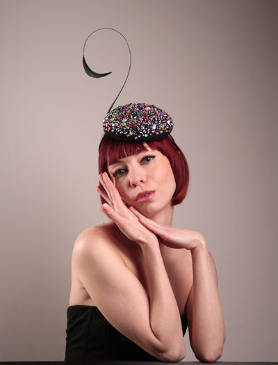 The model showcases the front of the Celestine pillbox fascinator. Its structured, button-shaped black base is drenched in a kaleidoscope of iridescent crystals—pink, aqua, violet, and gold, among others—each catching the light in motion. Crowned with a sweeping black quill that curves like a clef in mid-air. Designed by Merve Bayindir.