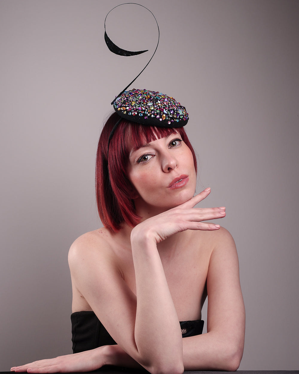 The model showcases the right front of the Celestine pillbox fascinator. Its structured, button-shaped black base is drenched in a kaleidoscope of iridescent crystals—pink, aqua, violet, and gold, among others—each catching the light in motion. Crowned with a sweeping black quill that curves like a clef in mid-air. Designed by Merve Bayindir.