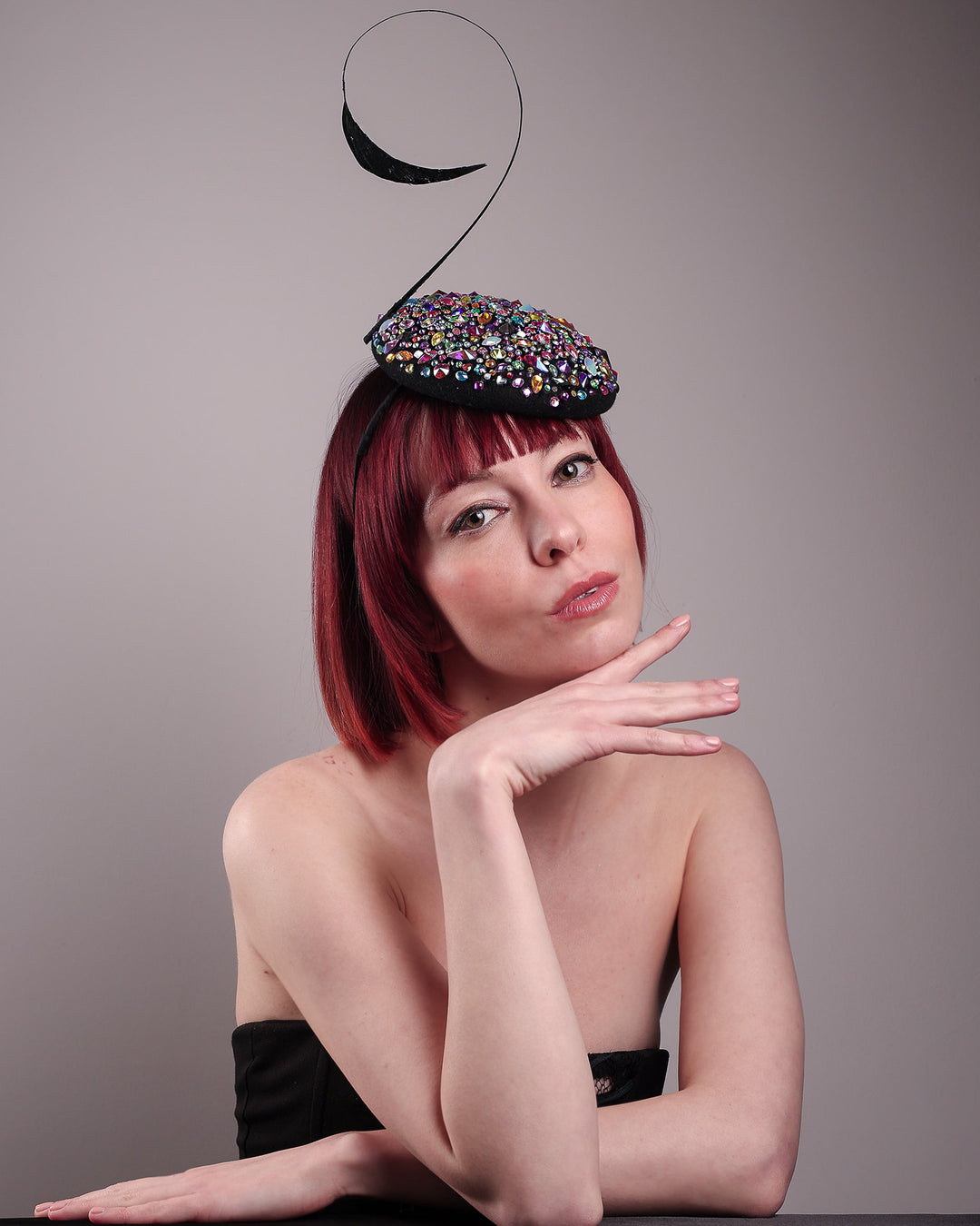 The model showcases the right front of the Celestine pillbox fascinator. Its structured, button-shaped black base is drenched in a kaleidoscope of iridescent crystals—pink, aqua, violet, and gold, among others—each catching the light in motion. Crowned with a sweeping black quill that curves like a clef in mid-air. Designed by Merve Bayindir.