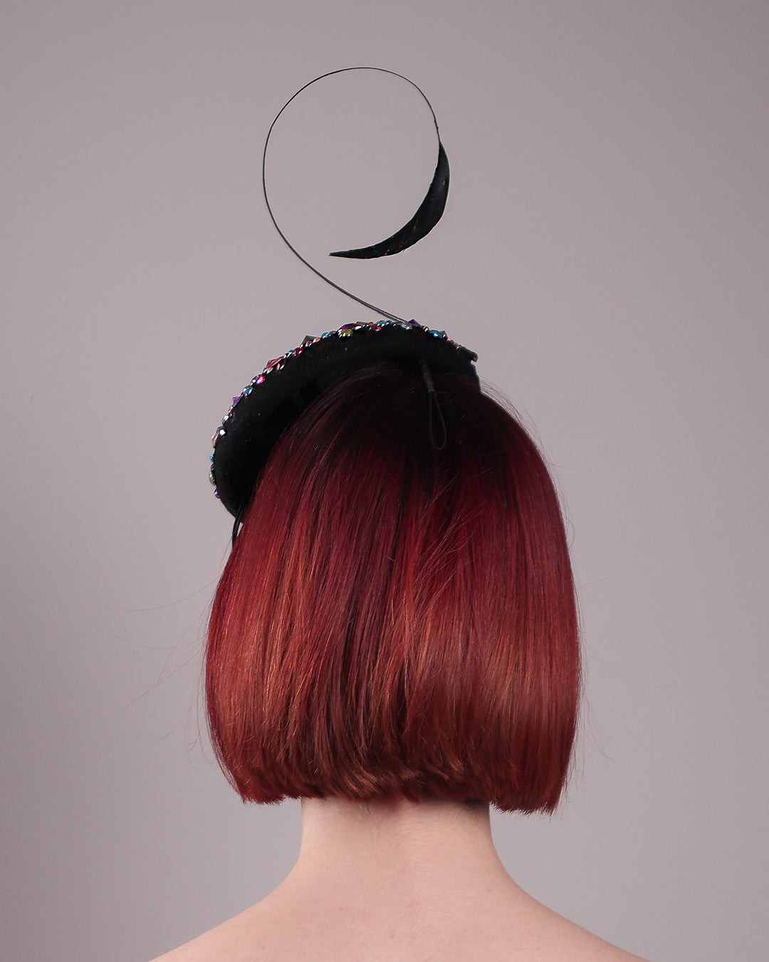 The model showcases the back of the Celestine pillbox fascinator. Its structured, button-shaped black base is drenched in a kaleidoscope of iridescent crystals—pink, aqua, violet, and gold, among others—each catching the light in motion. Crowned with a sweeping black quill that curves like a clef in mid-air. Designed by Merve Bayindir.