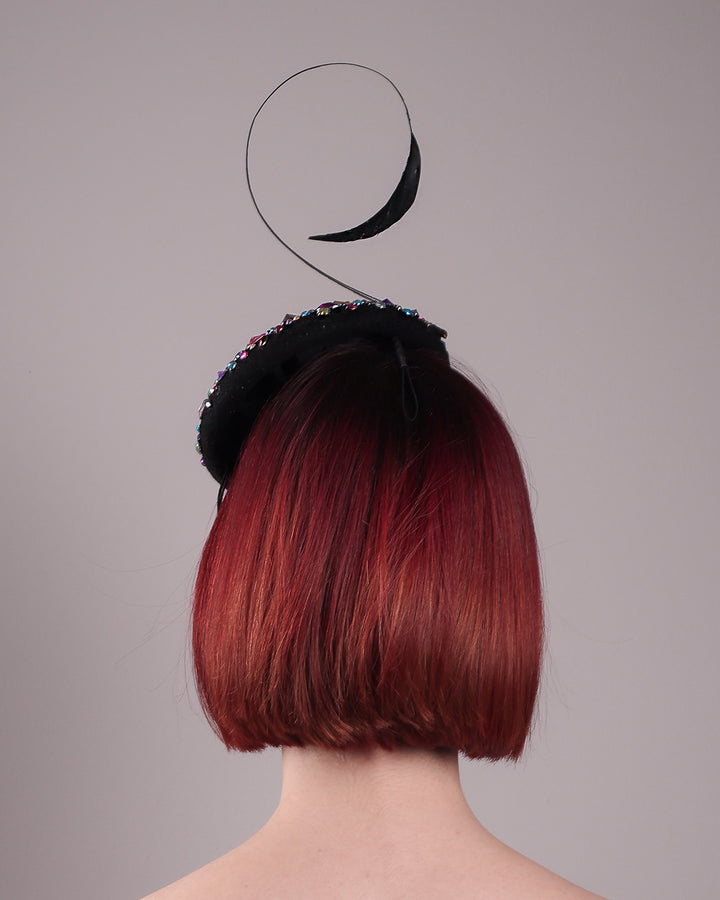 The model showcases the back of the Celestine pillbox fascinator. Its structured, button-shaped black base is drenched in a kaleidoscope of iridescent crystals—pink, aqua, violet, and gold, among others—each catching the light in motion. Crowned with a sweeping black quill that curves like a clef in mid-air. Designed by Merve Bayindir.